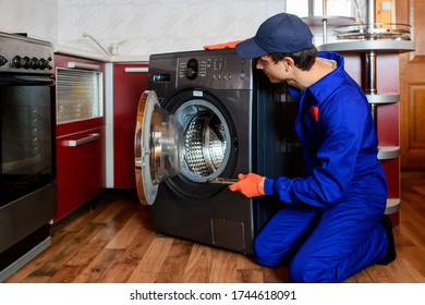 Young Man Plumber Handyman In Blue Overalls Repair/fix The Washing Machine In The Kitchen.