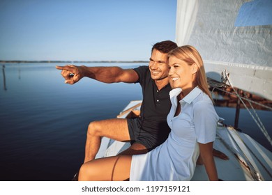 Young Man Pointing To Something On The Ocean Horizon To His Wife While Sitting On The Deck Of Their Boat Enjoying The Day Sailing