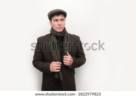 Young man posing in a brown winter coat with a wool scarf and flat cap, offering a thoughtful, confident fashion image. The guy is very serious, sad, doesn't want to grow up, to be responsible