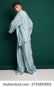Young man posing in striped loose fit shirt and wide pants with white sneakers against dark green background in studio. Fashion model with curly hair and casual outfit.