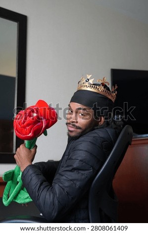 A young man posing with two large novelty roses while wearing a crown.