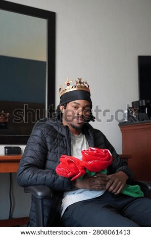 A young man posing with two large novelty roses while wearing a crown.