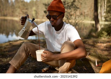 Young Man Pouring Coffee In Cup, Morning Breakfast On Open Air. Afro American Traveler Camping For First Time, New Experience To Rest With Tent. Nature, Leisure, No Rush, Peace And Calmness Concept