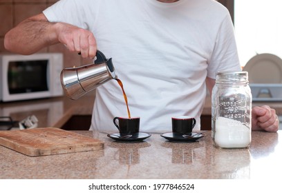 young man pouring freshly brewed coffee from a stainless steel coffee pot with a drop on a cup, and a pot of sugar with an unfocused background of a kitchen. falling coffee jet