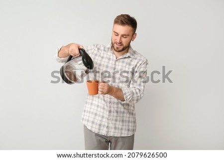 Young man pouring hot boiled water from electric kettle into cup on light background