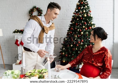 young man pouring water into glass near elegant and pregnant asian wife at romantic Christmas supper