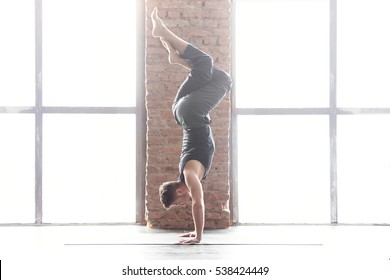 Young man practicing yoga in a urban background. Yogi handstand in pose shirshasana or Sirshasana with entwined legs near a picture window with red brick wall