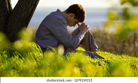 young man praying to God near a tree in the nature bowing his head to his knees with gratitude, male asks for help finding solace in faith, concept religion