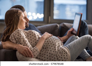 Young man and pregnant woman lying on sofa and using digital tablet
