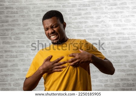 Young man presses hand to chest has heart attack suffers from unbearable pain closes eyes poses against grey brick wall background. People young age and problems with health concept, copy space.