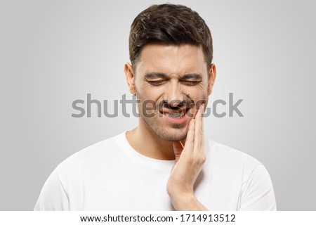 Young man pressing fingers to his jaw, trying to overcome severe toothache, eyes closed, isolated on gray background