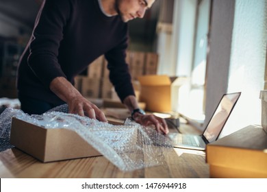 Young Man With A Product Looking At Laptop. Entrepreneur Verifying The Shipment Details Before Delivering To Customer.