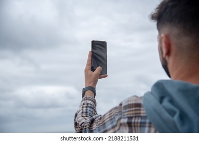 Young man in profile taking a picture of the landscape with his mobile phone on a cloudy day with the sky in the background.