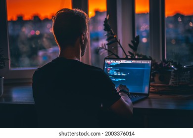 A Young Man Programmer Coding On A Laptop In The Dark With A View Of The Lights Of The Night City, Color Lighting In The Room. Coding And Hacking Concept