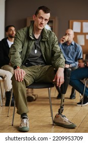 Young Man With Prosthetic Leg Sitting In Front Of Camera Against Counselor And Other Attendants Of PTSD Support Group Having Break