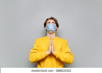 Young Man In A Protective Mask On His Face Prays And Looks Up On A Gray Wall Background. Guy Prays In A Medical Mask, Does Not Want To Get Coronavirus.