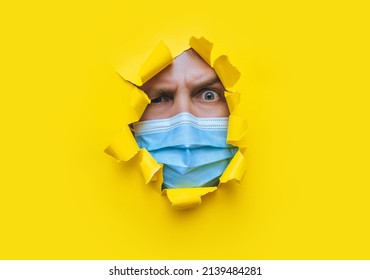 A Young Man In A Protective Medical Mask Looks In Surprise And Angrily With Curiosity Into A Broken Hole In Yellow Paper. The Concept Of Pandemic, Quarantine, Protection Against Coronavirus Covid-19.