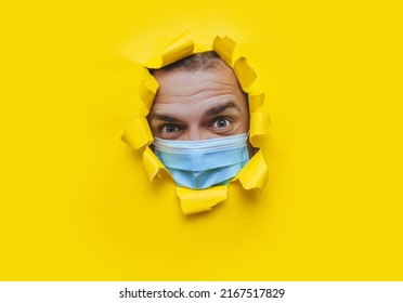 A Young Man In A Protective Medical Mask Looks In Surprise And Angrily With Curiosity Into A Broken Hole In Yellow Paper. The Concept Of Pandemic, Quarantine, Protection Against Coronavirus, Covid-19.