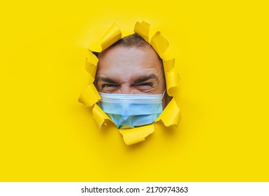 A Young Man In A Protective Medical Mask Looks In Surprise And Angrily With Curiosity Into A Broken Hole In Yellow Paper. The Concept Of Pandemic, Quarantine, Protection Against Coronavirus, Covid-19.