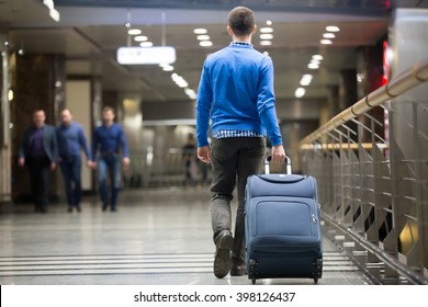 Young man pulling suitcase in modern airport terminal. Travelling guy wearing smart casual style clothes walking away with his luggage while waiting for transport. Rear view. Copy space
