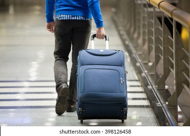 Young man pulling suitcase in modern airport terminal. Travelling guy wearing smart casual style clothes walking away with his luggage while waiting for transport. Rear view. Close-up