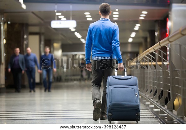 Young\
man pulling suitcase in modern airport terminal. Travelling guy\
wearing smart casual style clothes walking away with his luggage\
while waiting for transport. Rear view. Copy\
space