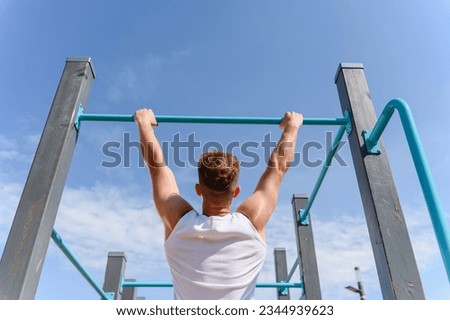 A young man pulls himself up on a horizontal bar on a street sports ground for a workout. View from the back. Blue sky in the background.