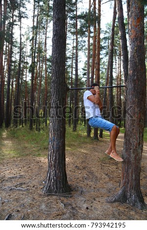 a young man pulls himself up on one arm on a horizontal bar in the woods