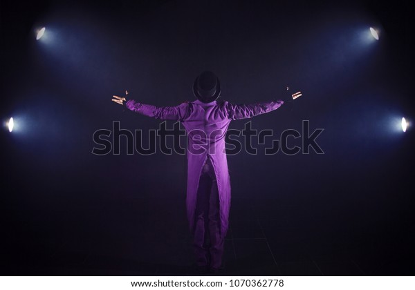Young man\
in purple suit standing on the background of the spotlight. Showman\
spreading hands, show begins. Back\
view