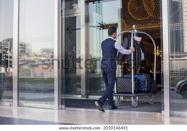 Young man\
pushes luggage cart with suitcases, bags and backpacks to the\
entrance. Metall baggage trailer with\
luggage