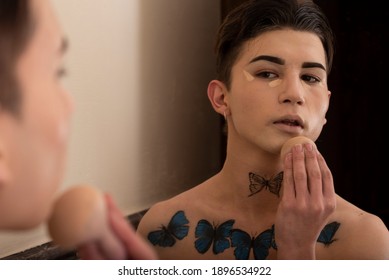 Young Man Put On Makeup In Front Of A Mirror With A Blue Butterfly Tattoo On His Chest. Warm Light And Selective Focus