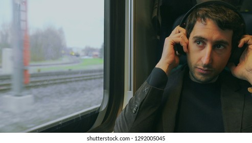 Young Man Putting Headphones While Riding Train And Looking Out Window