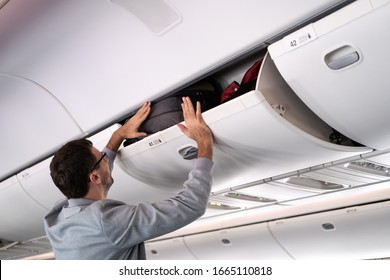 Young man putting luggage into overhead locker on airplane. Traveler placing carry on bag in overhead compartment in aircraft