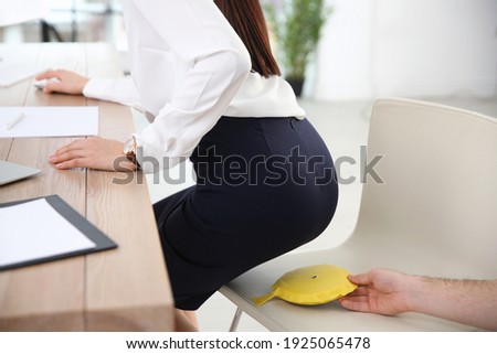 Young man putting whoopee cushion on chair while his colleague sitting down in office, closeup. Funny joke