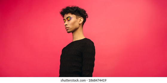 Young man with rainbow eye shadow makeup standing on red background with his eyes closed. Gender fluid man in black t-shirt.