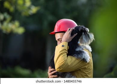 Young Man In Raincoat And With Helmet Holding Dog In Arms. Animal Rescue Team