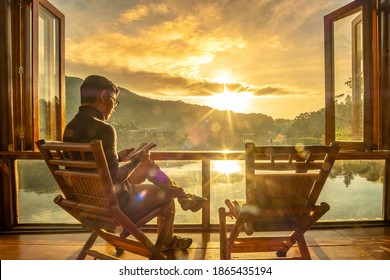 Young Man Reading Book Near Window And Watching Lake View At Coffee Shop In The Morning Sunrise, Ban Rak Thai Village, Mae Hong Son Province, Thailand. Travel Concept