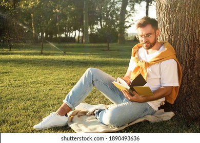 Young man reading book on green grass near tree in park - Powered by Shutterstock