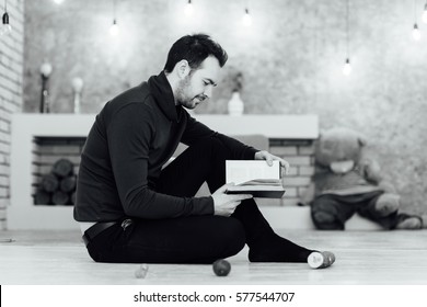 Young Man Reading A Book Seated On The Floor At Home. Black And White Photo