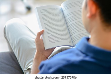 Young Man Reading Book While Sitting On The Couch, Over Shoulder View With Soft Focus