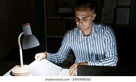 Young man reading documents in a dimly lit office late at night with laptop and desk lamp. - Powered by Shutterstock