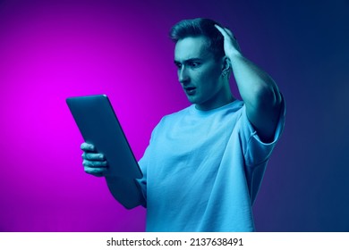 Young Man Reading News On Tablet With Shocked Expression Isolated On Gradient Pink Purple Background In Blue Neon. Looking Thoughtful. Concept Of Facial Expression, Fashion, Youth, Lifestyle, Emotions