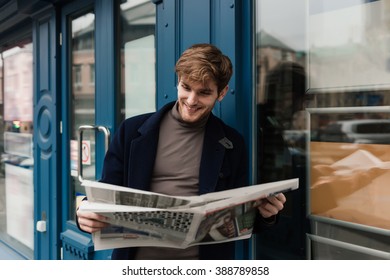 Young Man Reading Newspaper On The City Street, Leaning On Wall Of Modern Building.