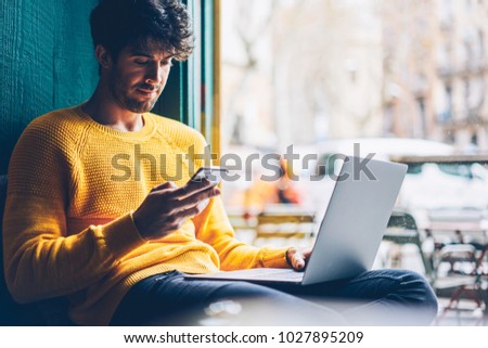 Young man reading sms message from friend on smartphone sitting with laptop device in stylish coworking space.Skilled student installing new application on mobile phone via free internet connection