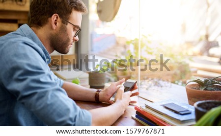 Young man reading a text message on his cellphone while sitting alone at a table by the window in a cafe 