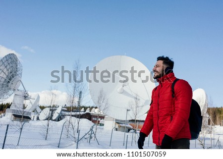 Young man in red jacket with backpack standing near the radio observatory with big satellite dishes or radio antennas in the forest in winter. Norway