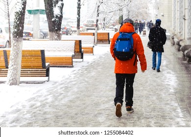 A young man in a red jacket with a blue backpack walks on a snow-covered sidewalk during a snowstorm, snowfall in the winter city.