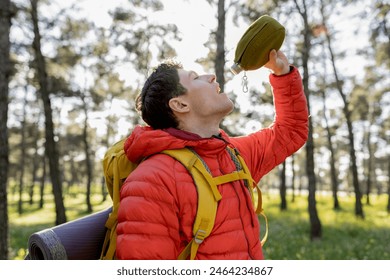 Young man in a red jacket drinking water from a canteen during a hike in the forest, staying hydrated. - Powered by Shutterstock