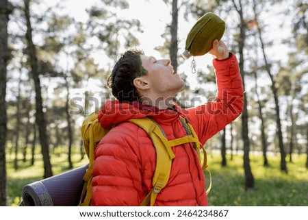 Young man in a red jacket drinking water from a canteen during a hike in the forest, staying hydrated.