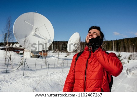 Young man in red jacket talking on the smart phone while standing near the radio observatory with big satellite dishes and radio antennas in the forest on bright winter day. Norway